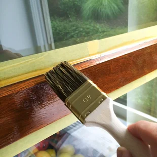Klebebänder - Mit Hilfe eines Pinsels wird Holzlasur auf einen Holzfensterrahmen aufgetragen, die Glasscheibe wurde mit Malercrep abgeklebt.