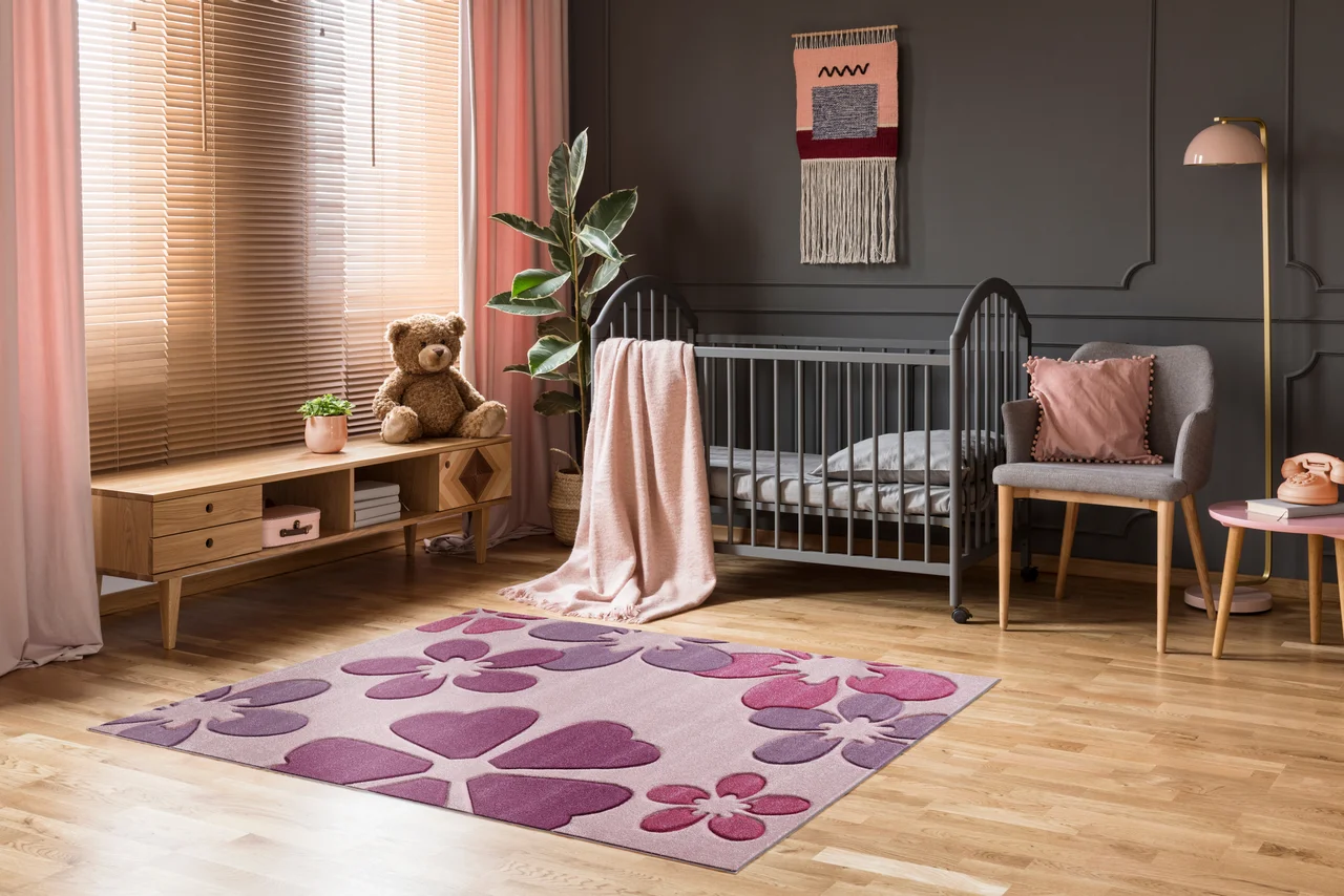 Illustration - Ihr Experte für schönes Wohnen - Rosa Kinderteppich mit Blumenmuster liegt im Kinderzimmer vor dem Kinderbett.